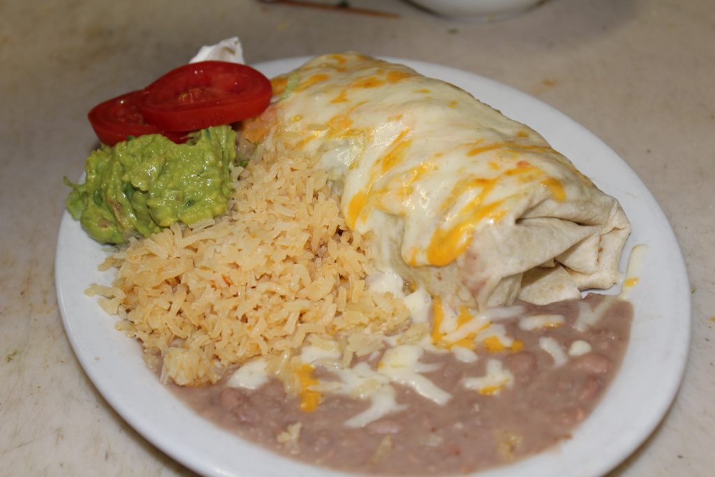 A burrito covered in cheese on a plate with rice, beans, guacamole, and tomato slices.