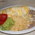 A burrito covered in cheese on a plate with rice, beans, guacamole, and tomato slices.